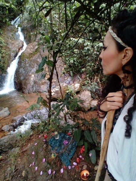 A woman looks away towards a rocky waterfall flowing into a small stream. On the rocky riverbank beside her are small offerings as part of a gratitude ritual to the water.